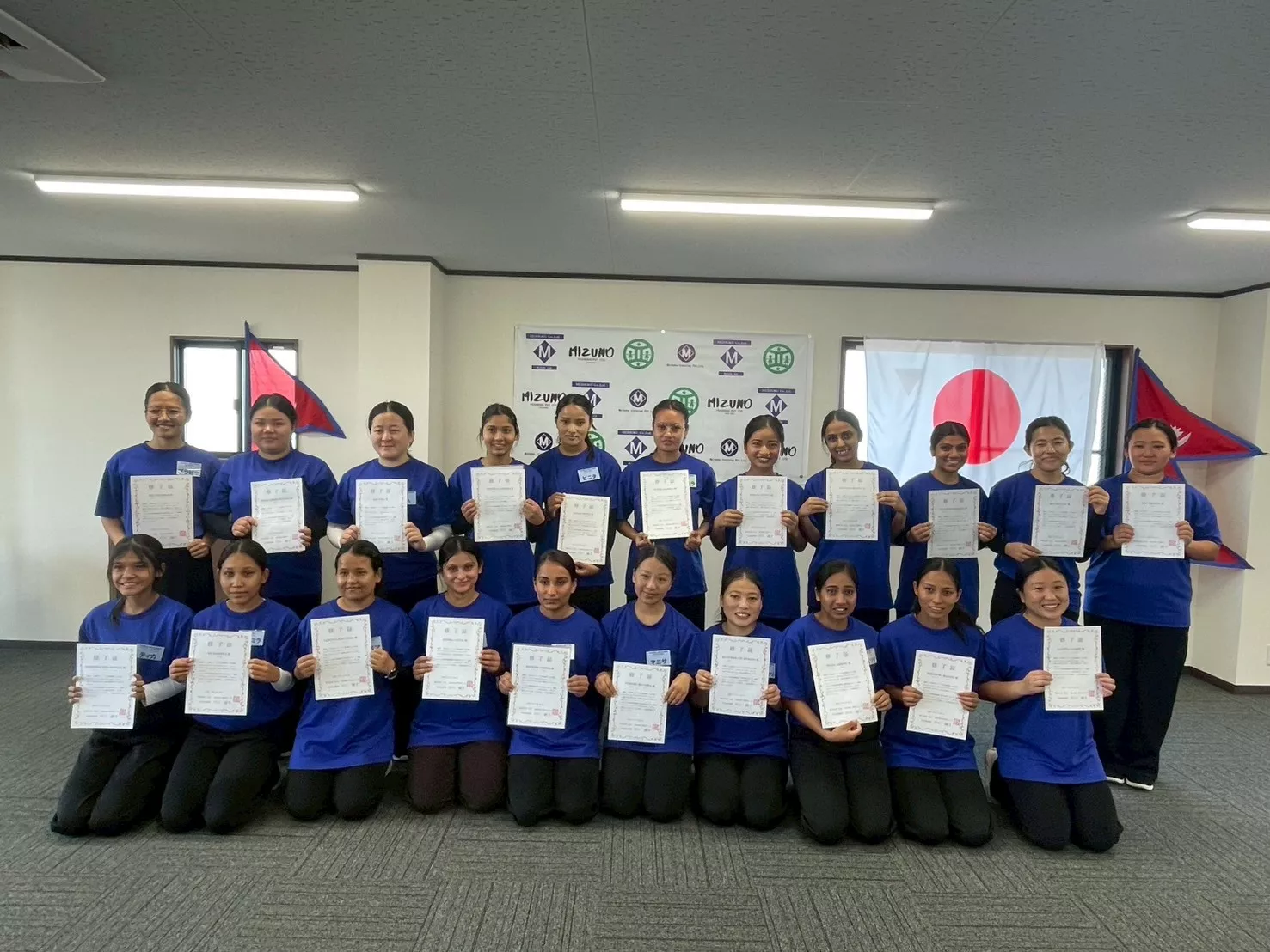 A group of women in blue shirts proudly hold certificates, standing in two rows. Behind them are Japanese and Nepalese flags and a banner with logos. The mood is joyful.