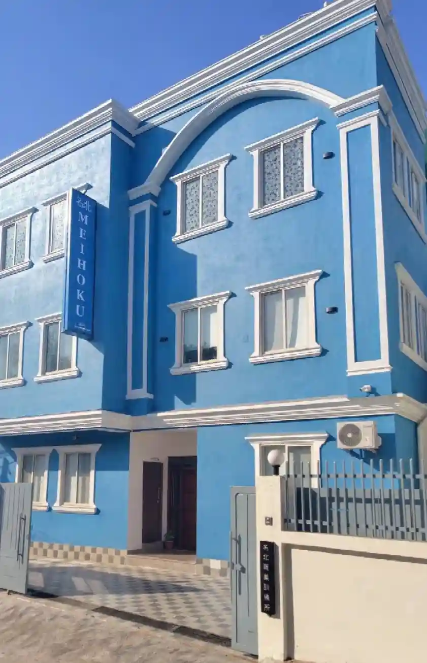Blue building with white trim, featuring arched windows and a "Meihoku" sign. Open gates and a small courtyard create a welcoming atmosphere.