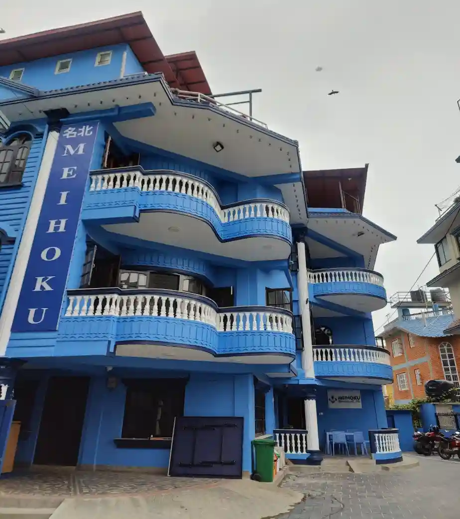 A vibrant blue building with "MEIHOKU" vertically on a sign, features ornate balconies and white railings. A bird flies above, creating a dynamic urban feel.