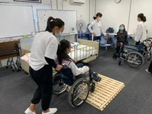People in a classroom are participating in a wheelchair training exercise. A woman assists a child over a wooden ramp. Others observe, creating a supportive and focused environment.