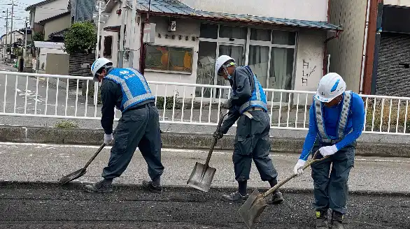 Three construction workers in blue uniforms and helmets use shovels to level asphalt on a road in a residential area.