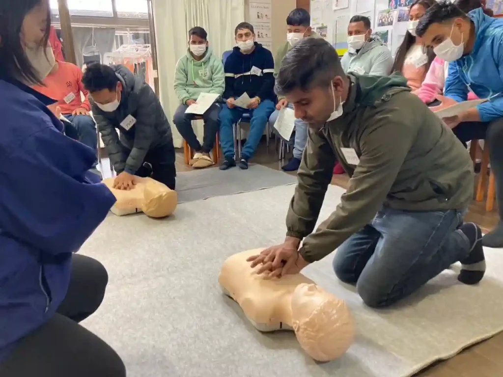People in a classroom participate in a CPR training session, practicing chest compressions on mannequins. Others observe, wearing masks and holding notes.