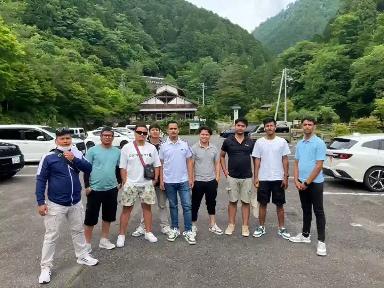 A group of nine people stands in a parking lot surrounded by lush green mountains. They appear relaxed and happy, with a wooden building and parked cars in the background.