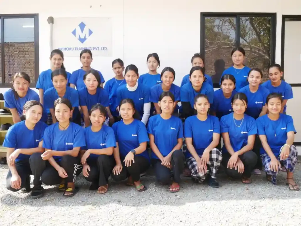 A group of 23 young women in blue shirts and black pants pose together in two rows outside Meihoku building . They appear focused, conveying teamwork.