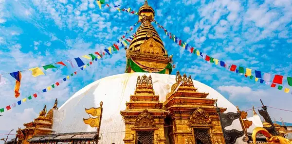 Swayambhunath stupa with colorful prayer flags stretches into a bright blue sky with scattered clouds. The scene feels vibrant and culturally rich.