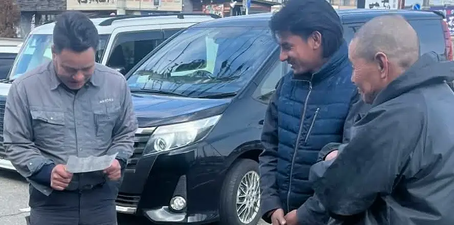 Three people stand in front of parked cars. One person on the left reads a paper, while the two on the right smile, creating a friendly, relaxed atmosphere.