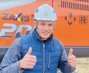 Man in a blue vest and safety helmet gives a thumbs up, standing beside an orange construction vehicle. He appears cheerful and confident.
