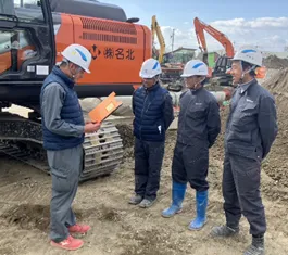 Four construction workers in helmets and jackets discuss plans near an orange excavator on a construction site. The mood is professional and focused.