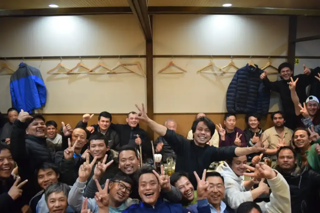 A large group of people in a cozy room celebrate joyously, making peace signs and smiling. Coats hang on hooks behind them, conveying warmth and camaraderie.