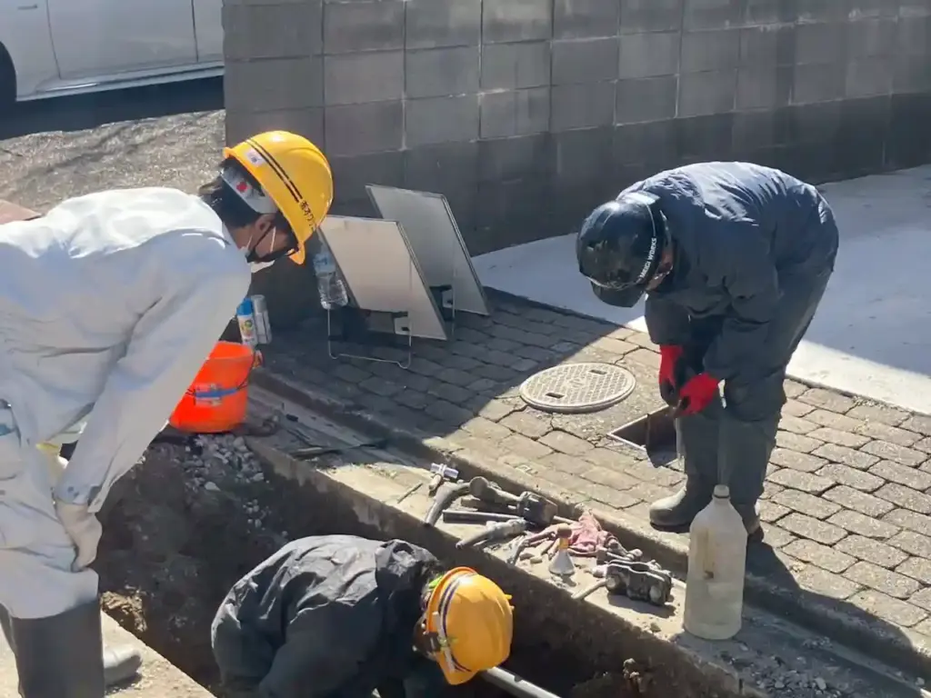 Three construction workers wearing protective gear and helmets work in a trench on a sunny day. Nearby, an orange bucket and tools are visible.
