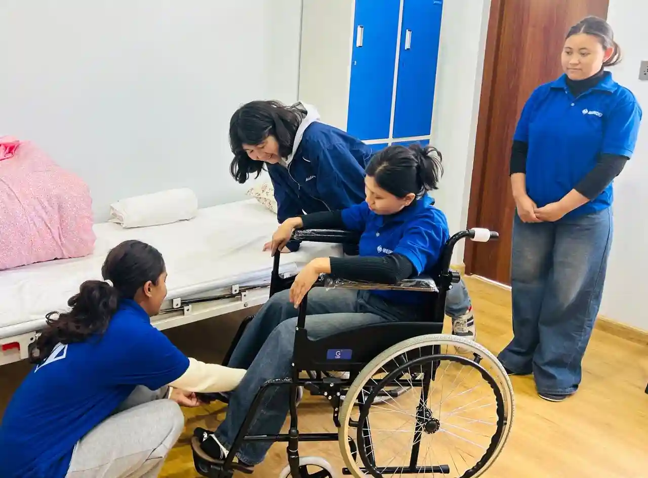 A woman is sitting in a wheelchair with three people assisting her in a bright room with wooden floors. A bed with pink sheets is nearby, suggesting a care setting. The mood is supportive and focused.