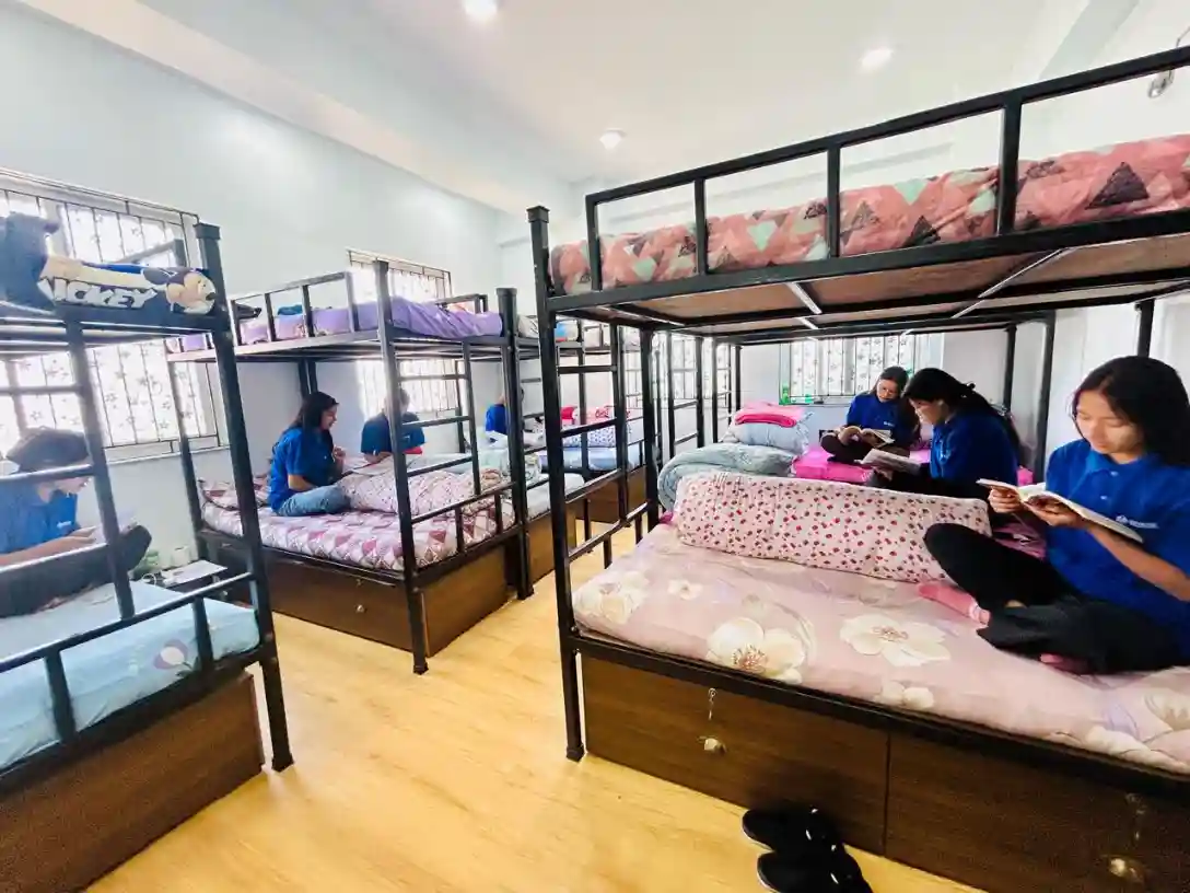 A dorm room with bunk beds, featuring five people in blue shirts reading or relaxing. The room is bright with wooden floors and colorful bedding.