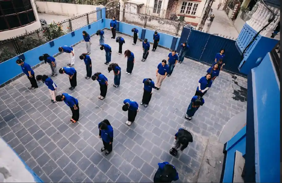 A group of people in blue uniforms bend forward in unison in a courtyard with blue walls and gray tiles, conveying discipline and order.