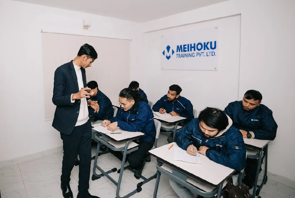 A teacher instructs attentive students seated at desks, taking notes in a classroom with "Meihoku Training Pvt. Ltd." sign on the wall.