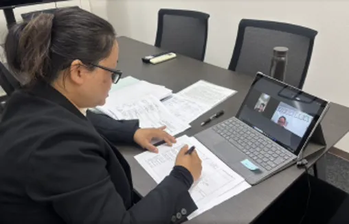 A woman in a black blazer, with hair tied back, is on a video call via laptop, surrounded by documents, in an office setting. She appears focused.