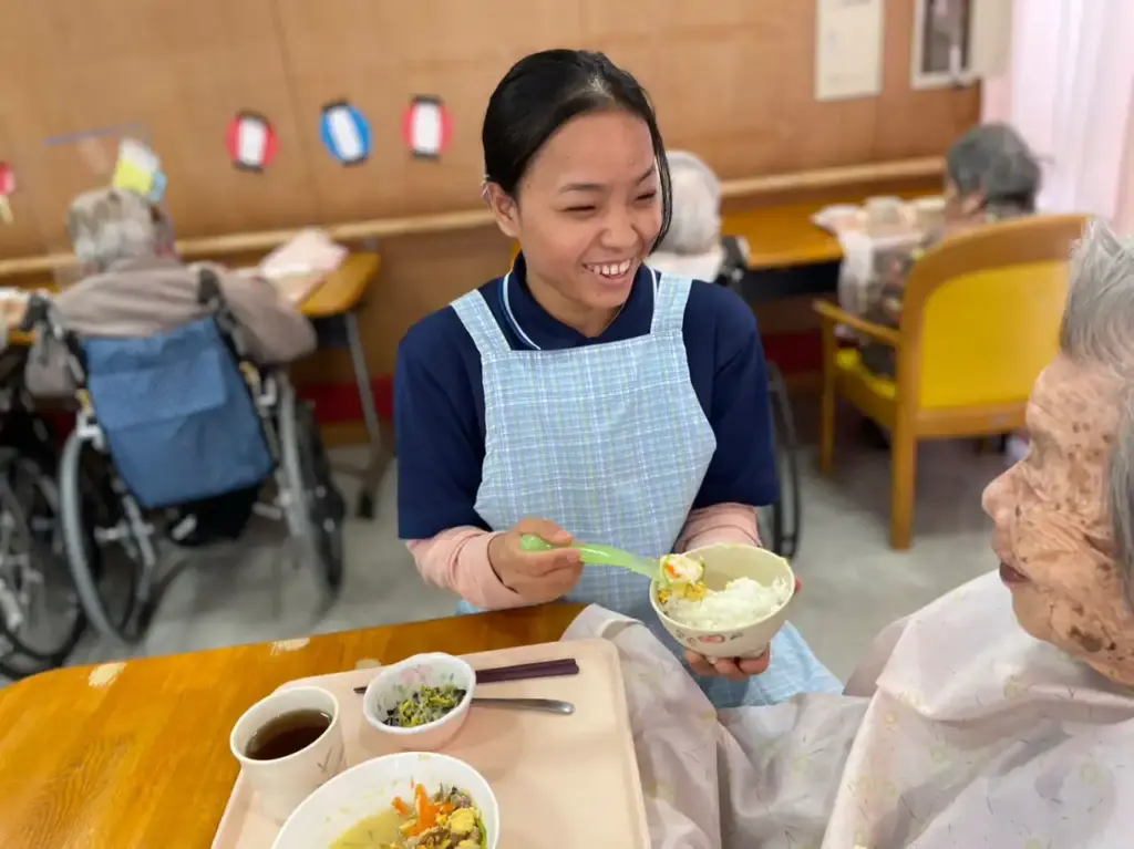 A caregiver in a blue apron smiles warmly while feeding an elderly person seated at a table with a tray of food. The setting is a cozy dining area with other seniors in the background, creating a caring and joyful atmosphere.