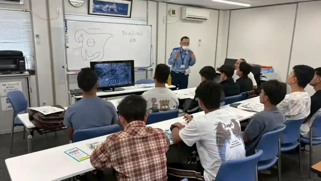 A classroom setting with a police officer instructing a group of students seated at tables. A monitor displays a map; the whiteboard has a sketch.