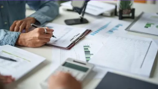 Hands holding a pen over financial charts on a desk, surrounded by papers, a calculator, and a potted plant, conveying a focused work environment.