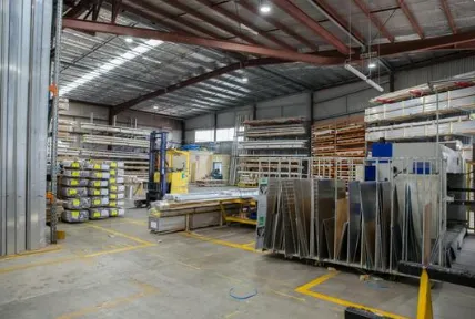 Spacious warehouse interior with stacked metal sheets and wooden planks. High shelves line the walls, yellow forklift visible, well-lit with overhead lights.