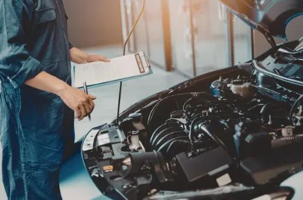 Mechanic in blue coveralls holding a clipboard inspects a car engine with the hood open in a well-lit garage, conveying a professional, focused atmosphere.