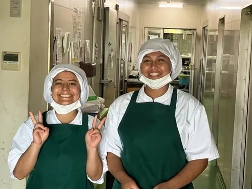 Two people wearing white uniforms and green aprons stand smiling in a corridor. One makes a peace sign. The setting appears to be a kitchen area.