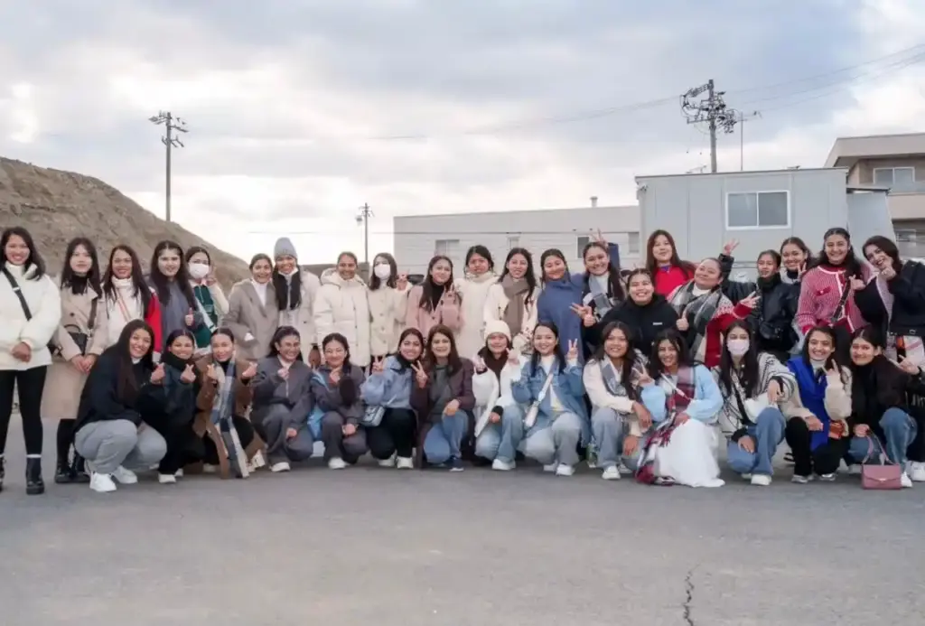 A group of young women in winter clothing pose outdoors on a cloudy day, smiling and making peace signs, conveying a joyful and united atmosphere.
